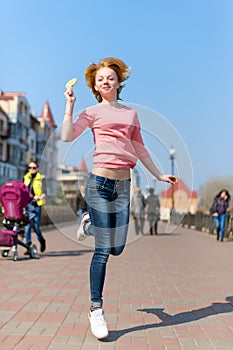 Redhead beautiful young woman jumping high in air over blue sky holding colorful lollipop. Pretty girl having fun outdoors.