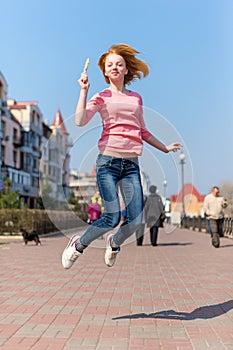 Redhead beautiful young woman jumping high in air over blue sky holding colorful lollipop. Pretty girl having fun outdoors.