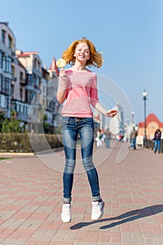 Redhead beautiful young woman jumping high in air over blue sky holding colorful lollipop. Pretty girl having fun outdoors.