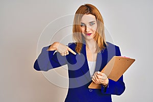 Redhead business caucasian woman holding clipboard over isolated background with surprise face pointing finger to himself