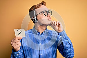 Redhead call center agent man working using headset holding reminder with question mark serious face thinking about question with