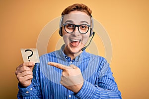 Redhead call center agent man working using headset holding reminder with question mark Smiling happy pointing with hand and