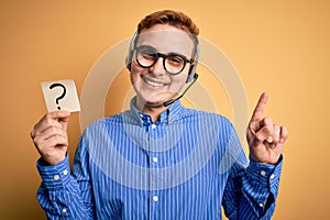 Redhead call center agent man working using headset holding reminder with question mark surprised with an idea or question