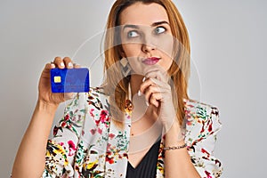 Redhead caucasian business woman holding credit card over isolated background serious face thinking about question, very confused