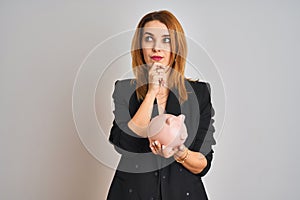 Redhead caucasian business woman holding piggy bank over isolated background serious face thinking about question, very confused