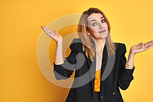 Redhead caucasian business woman over yellow isolated background clueless and confused expression with arms and hands raised