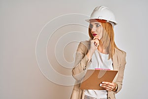 Redhead caucasian business woman wearing safety helmet and holding clipboard serious face thinking about question, very confused