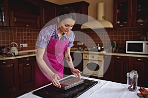 Redhead confectioner in pink apron holding chocolate molds full of liquid heated chocolate mass and putting it on a marble surface