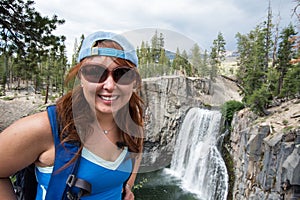 Redhead female hiker wearing a ballcap poses at Rainbow Falls in California along the John Muir Trail