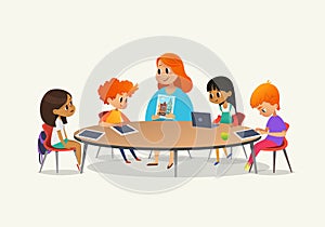 Redhead female teacher showing picture to children sitting around round table at class with laptop and tablet pc. Kids