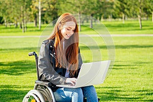 Redhead ginger freelancer woman sitting in wheelchair and working by laptop in summer park