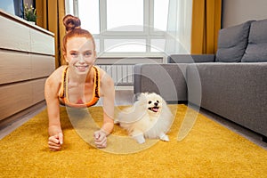 Redhead ginger girl lying down with white small dog spitz on the carpet and looking at camera