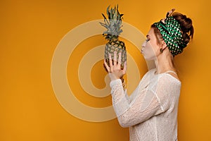 Redhead girl with a bright scarf on head is standing on an orange background in profile, holding a pineapple and