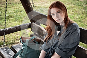 Redhead girl in a denim shirt sits with a backpack on a wooden swing bench and looks into the frame.