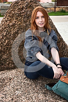 Redhead girl with freckles and blue eyes in a denim shirt with headphones on her neck sits on a stone and looks into the