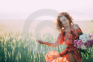 Redhead girl in a red dress with a bouquet of peonies dancing joyful in a wheat field in summer at sunset. Soft focus