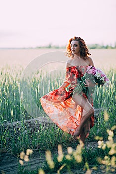 Redhead girl in a red dress with a bouquet of peonies dancing joyful in a wheat field in summer at sunset. Soft focus