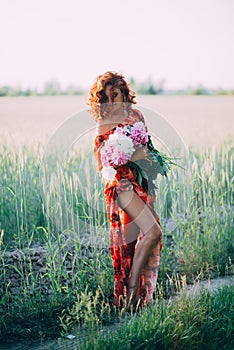 Redhead girl in a red dress with a bouquet of peonies dancing joyful in a wheat field in summer at sunset. Vertical
