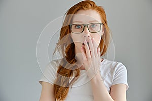 Redhead girl in a white T-shirt and glasses stands on a gray background, looks surprised into the frame, covering mouth