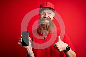 Redhead Irish delivery man with beard holding smartphone showing app on screen happy with big smile doing ok sign, thumb up with