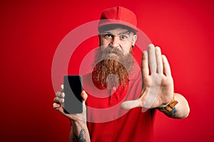 Redhead Irish delivery man with beard holding smartphone showing app on screen with open hand doing stop sign with serious and