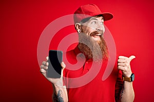 Redhead Irish delivery man with beard holding smartphone showing app on screen pointing and showing with thumb up to the side with