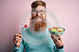 Redhead Irish healthy man with beard eating vegetarian green salad over pink background with a happy face standing and smiling