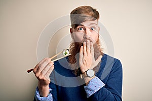 Redhead Irish man with beard eating green maki sushi using chopsticks over yellow background cover mouth with hand shocked with