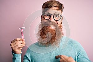 Redhead Irish man with beard holding beauty razor for shaving and skin care over pink background with surprise face pointing