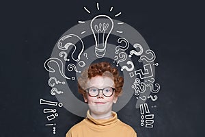 Redhead kid with lightbulb and question marks on blackboard background. Brainstorming and idea concept