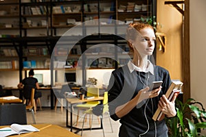 Redhead lady student posing indoors in library holding books istening music with earphones using mobile phone