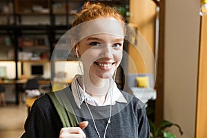Redhead lady student posing indoors in library istening music with earphones