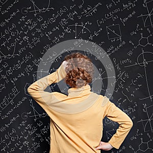 Redhead little boy thinking and looking at blackboard background with science formulas