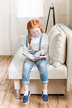 Redhead little girl sitting on sofa and reading book