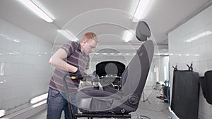 Redhead man is hoovering black car seat in a room of auto-service during cleaning, car washing