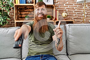 Redhead man with long beard holding television remote control smiling happy pointing with hand and finger to the side