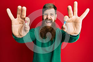 Redhead man with long beard holding two different pills smiling and laughing hard out loud because funny crazy joke