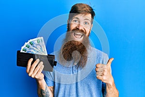 Redhead man with long beard holding wallet with south african rand banknotes pointing thumb up to the side smiling happy with open