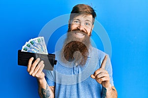 Redhead man with long beard holding wallet with south african rand banknotes smiling happy pointing with hand and finger to the