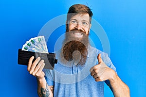 Redhead man with long beard holding wallet with south african rand banknotes smiling happy and positive, thumb up doing excellent