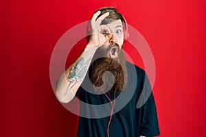 Redhead man with long beard listening to music using headphones doing ok gesture shocked with surprised face, eye looking through