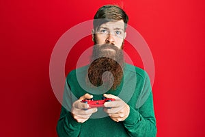 Redhead man with long beard playing video game holding controller relaxed with serious expression on face