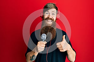 Redhead man with long beard singing song using microphone and wearing headphones smiling happy and positive, thumb up doing