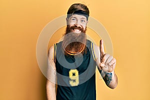 Redhead man with long beard wearing basketball uniform showing and pointing up with finger number one while smiling confident and