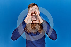 Redhead man with long beard wearing casual blue sweater over blue background shouting angry out loud with hands over mouth