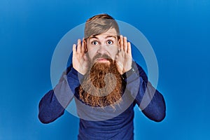 Redhead man with long beard wearing casual blue sweater over blue background trying to hear both hands on ear gesture, curious for