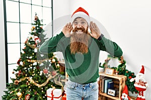 Redhead man with long beard wearing christmas hat by christmas tree trying to hear both hands on ear gesture, curious for gossip