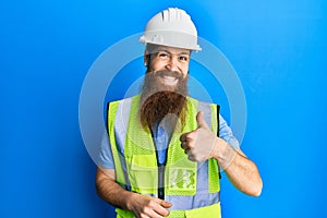 Redhead man with long beard wearing safety helmet and reflective jacket doing happy thumbs up gesture with hand