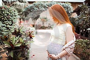 Redhead pregnant girl stroking her belly. On the background of the yard with plants. Summer