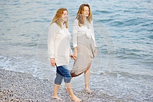 Redhead senior mother and her adult beautiful pregnant daughter are walking together on the sea shore